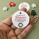 Etichetta tonda per bomboniere Natale, con grafica fiocco rosso e agrifoglio. Tag battesimo natalizio